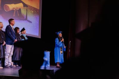 Ceremonia de graduación en el escenario; un graduado con toga azul habla por un micrófono. Cuatro profesores de pie a la izquierda, con la imagen de un diploma proyectada detrás.