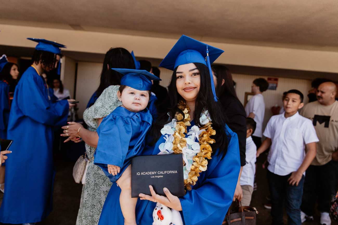 Joven con toga azul de graduación sostiene a un bebé con toga y birrete a juego, sonriendo orgullosa con un diploma. Ambiente festivo con multitud.