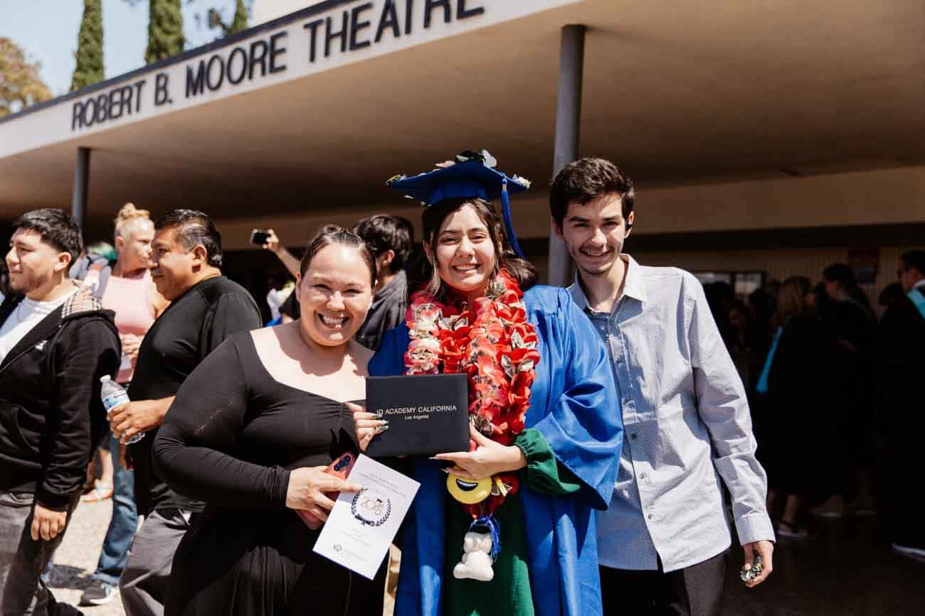 Una alegre graduada con toga y birrete azul sostiene un diploma, rodeada de sonrientes seguidores a las puertas del teatro Robert B. Moore.