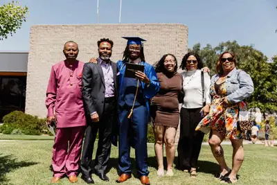 Un graduado con toga y birrete azul sostiene un diploma, rodeado de cinco familiares sonrientes en un día soleado, transmitiendo orgullo y celebración.